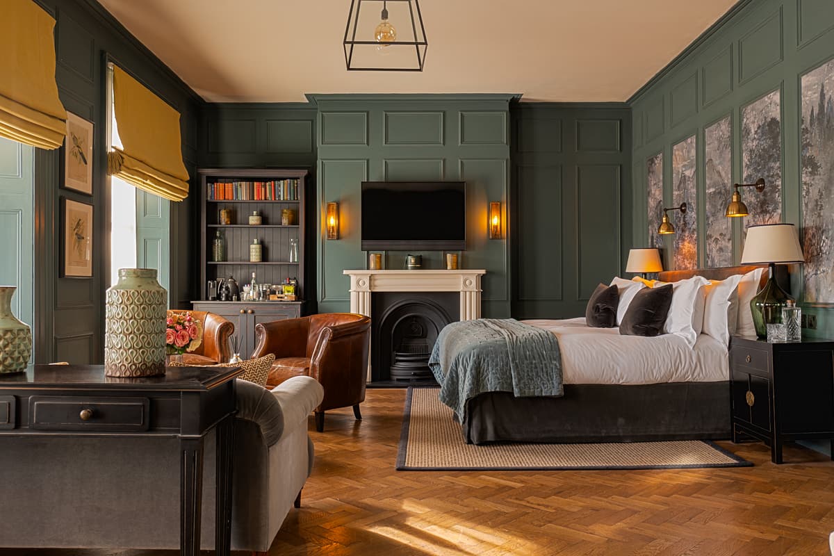 Hotel bedroom with dark green panelled walls, mustard blinds, a bed with a grey-green throw, a fireplace and a tan leather armchair on a parquet floor.