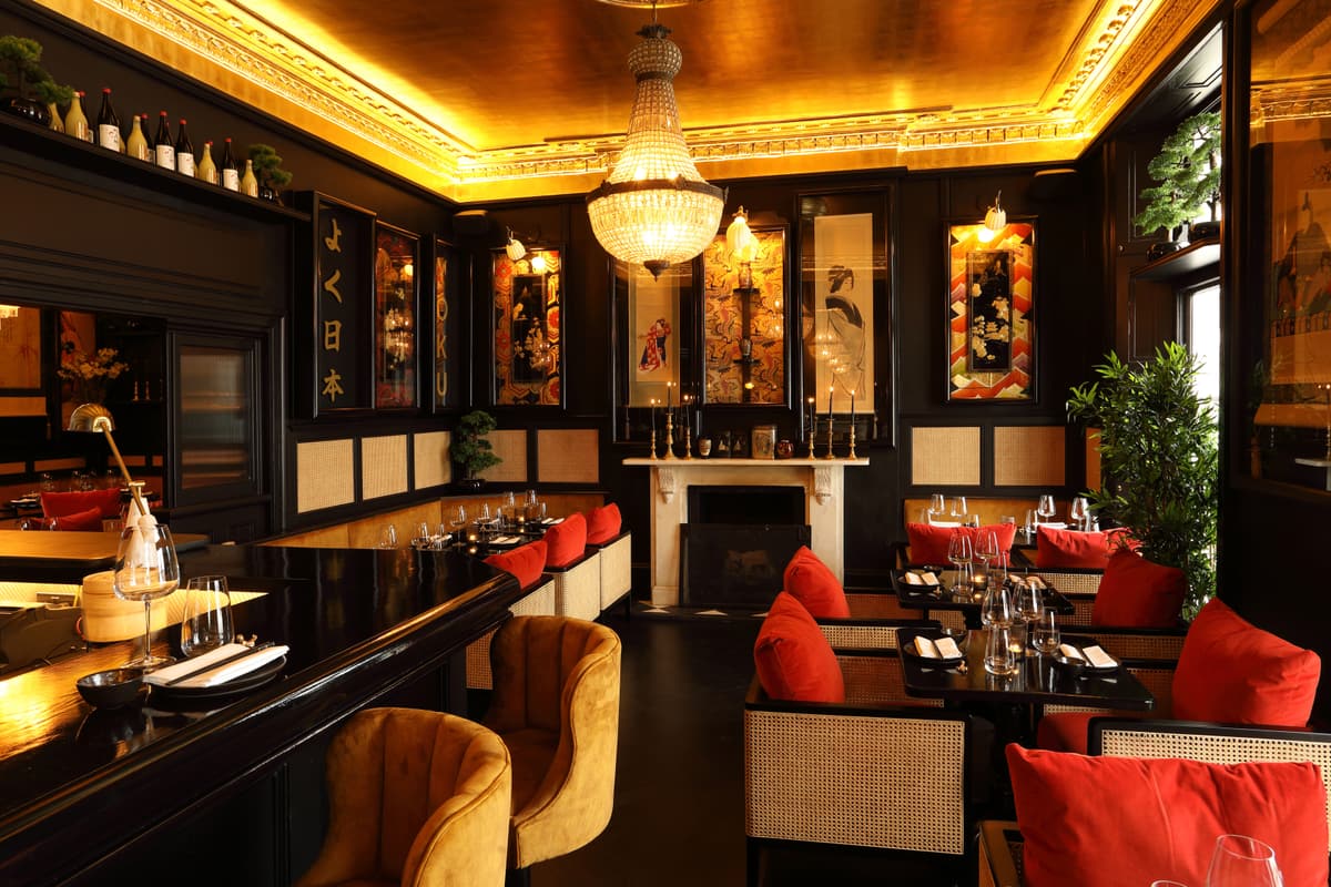 Dining room with a gold ceiling, a crystal chandelier, black panelled walls hung with Japanese prints and tables set beside red cushioned chairs.
