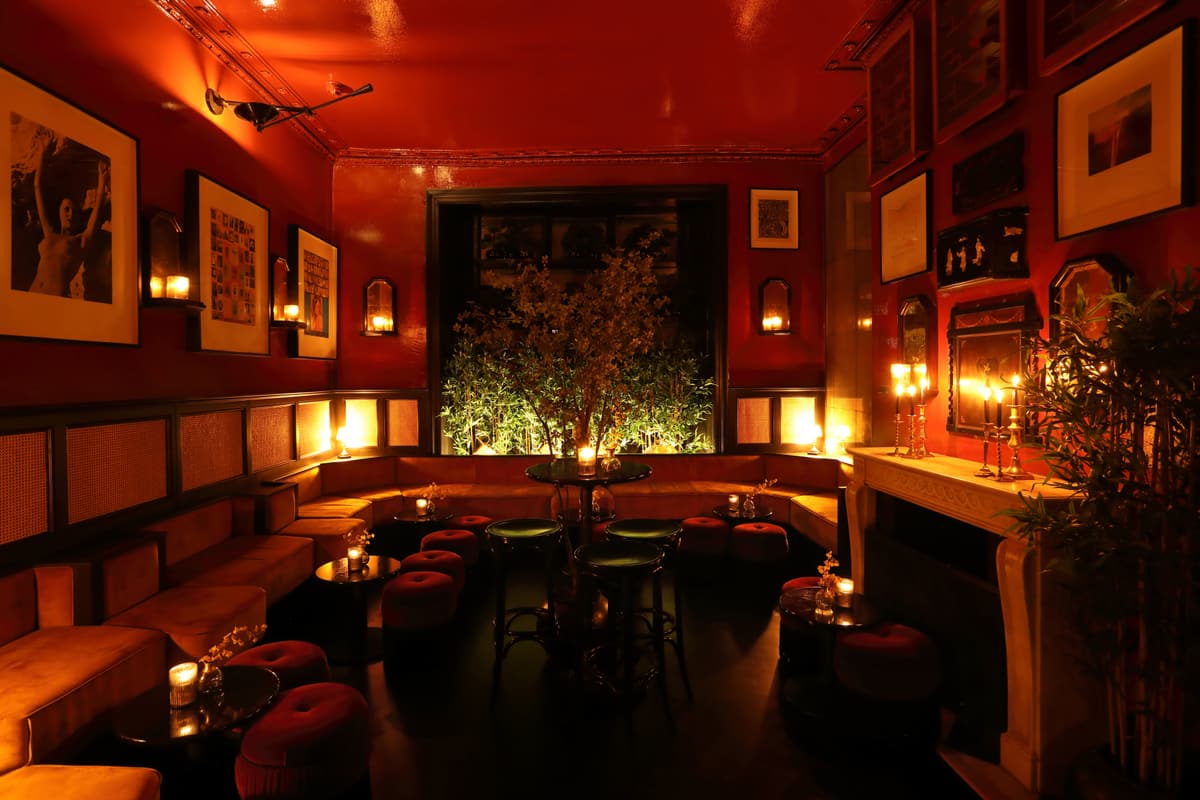 Lounge with red lacquered walls and ceiling, framed pictures, low velvet stools and banquette seating lit by candles.