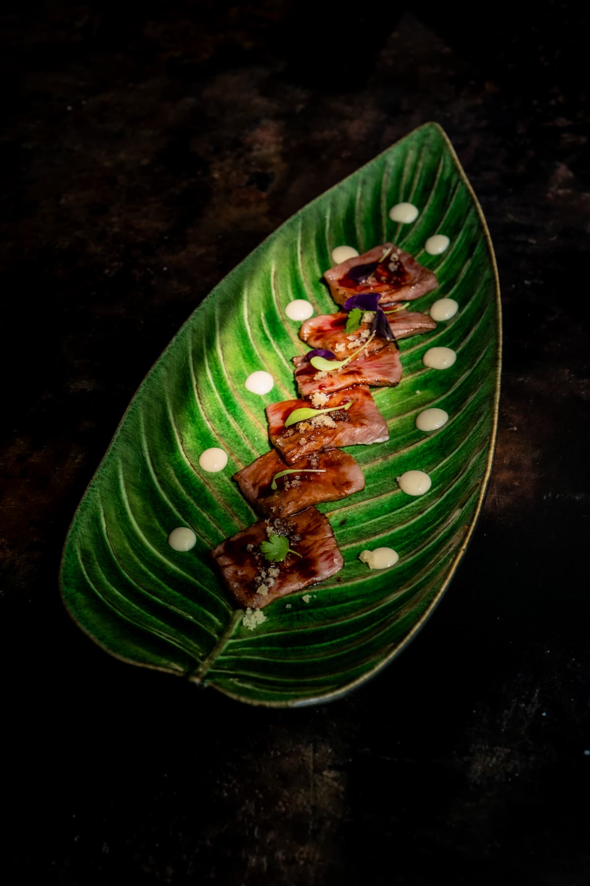 Seared slices garnished with small leaves and dots of sauce, arranged along a green leaf-shaped plate on a dark table.