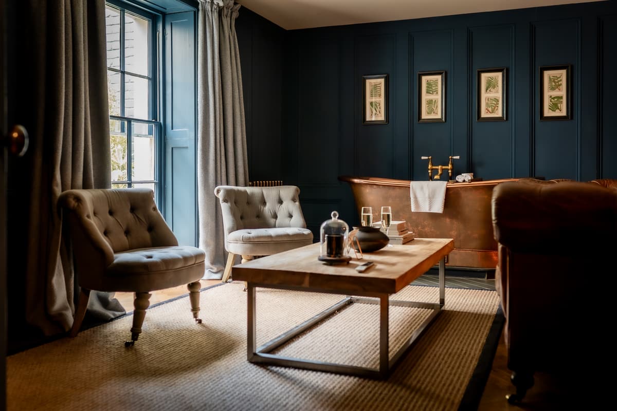 Sitting area in a dark blue panelled room with two buttoned armchairs, a wooden coffee table and a copper bathtub beside a sash window.