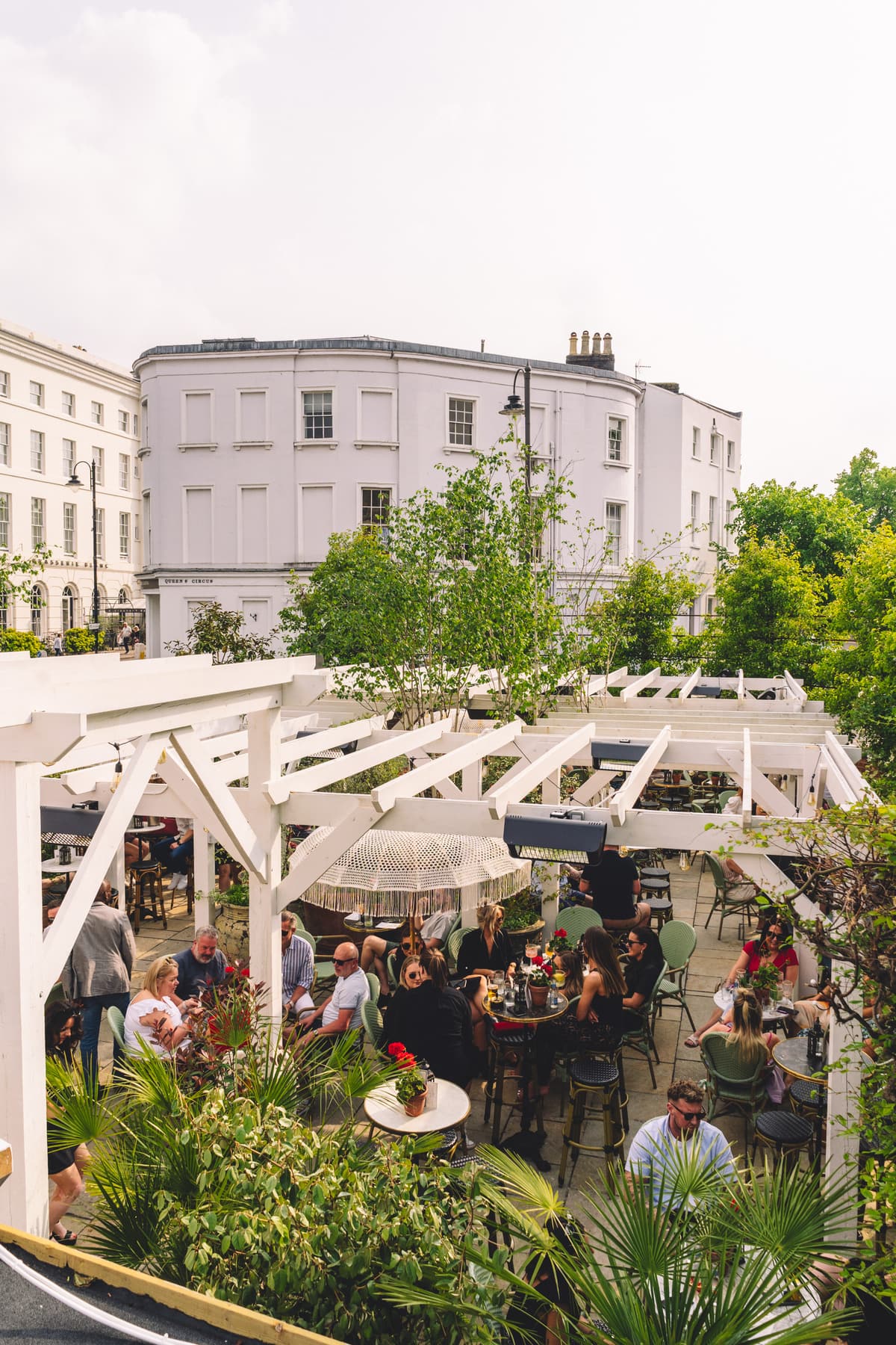 People eating and drinking at tables under white timber pergolas on a raised terrace, with planting in the foreground and white Regency buildings behind.